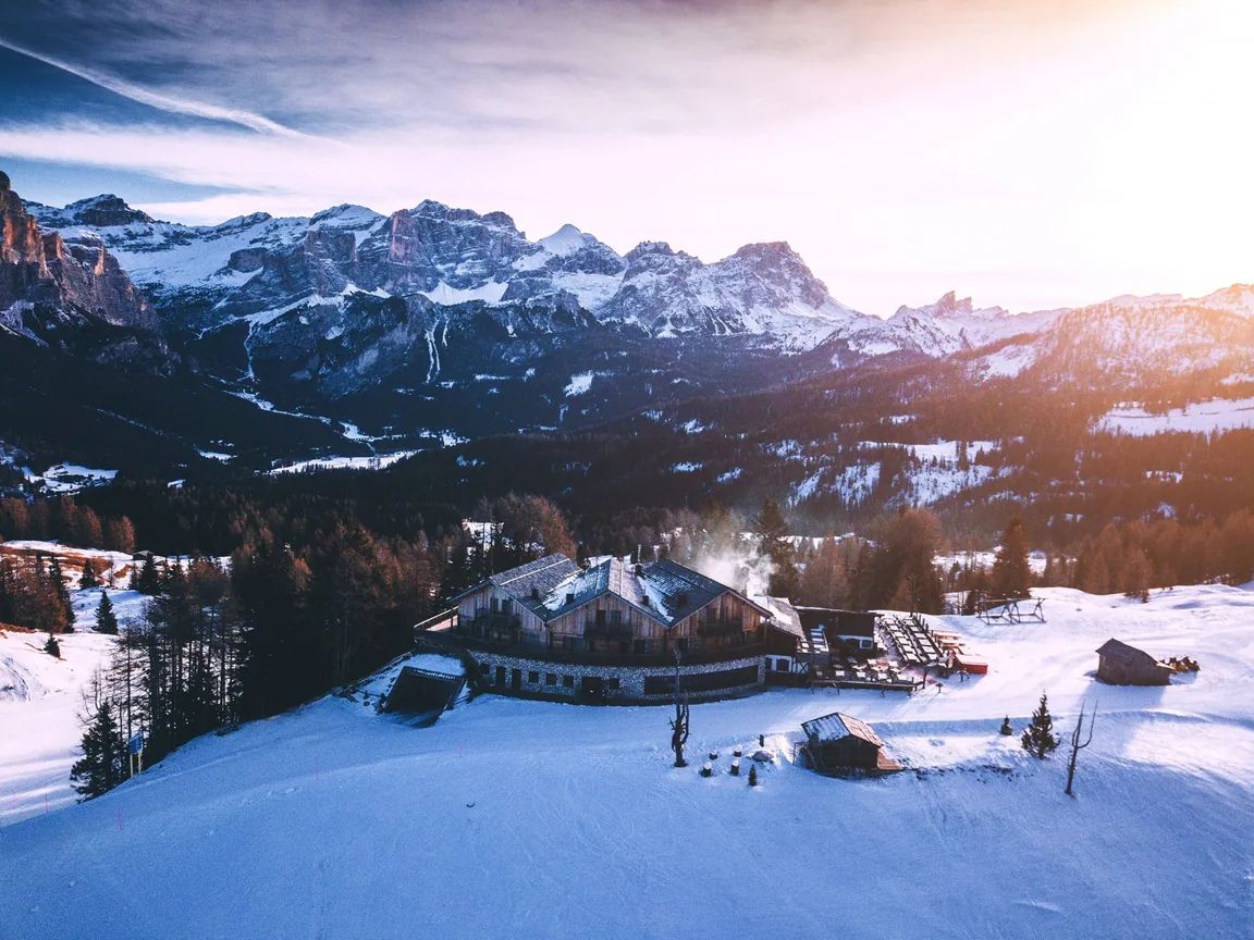 A timber mountain building on a snowy slope at sunset, with Dolomite peaks behind