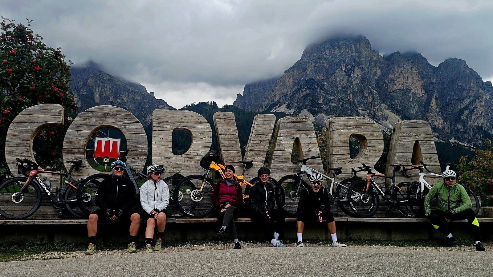 Cyclists in front of the wooden Corvara sign beneath cloudy mountains