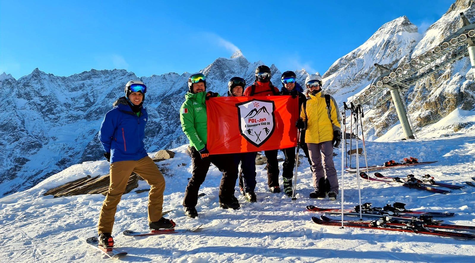 A group on a summit with a Pol-Ski flag and snowy ridges behind